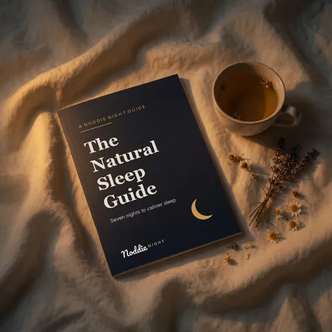 The Natural Sleep Guide, a navy booklet from Noddie Night, on linen beside a cup of chamomile tea.